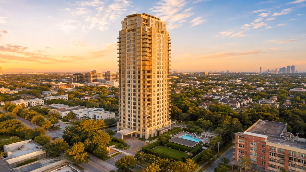 Aerial photograph of Montebello in low golden light: a cream-colored 30-story tower with recessed balconies stands at the centre of the frame, its sunlit side glowing warm, above a porte-cochere entrance, with a swimming pool, poolside pavilion, lawn and clipped hedges in the grounds beside it. Low white and brick buildings and tree-lined streets surround it, a red-brick mid-rise sits at the right edge, mid-rise office blocks stand on the horizon to the left and a cluster of high-rises on the horizon to the far right, under a sky of gold-lit and grey clouds.