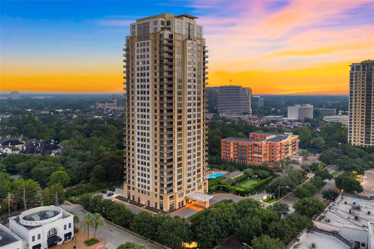 Aerial view at sunset of Montebello, a cream-colored 30-story tower with recessed balconies rising above a lit porte-cochere entrance, a swimming pool, lawn and putting green in the grounds beside it, low-rise brick buildings and tree-lined streets beyond, and an orange and pink sky over the horizon.